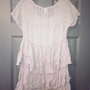May include: A light purple dress with a lace bodice and short sleeves. The dress features a tiered, ruffled skirt. The garment is hanging on a black hanger against a dark gray door.