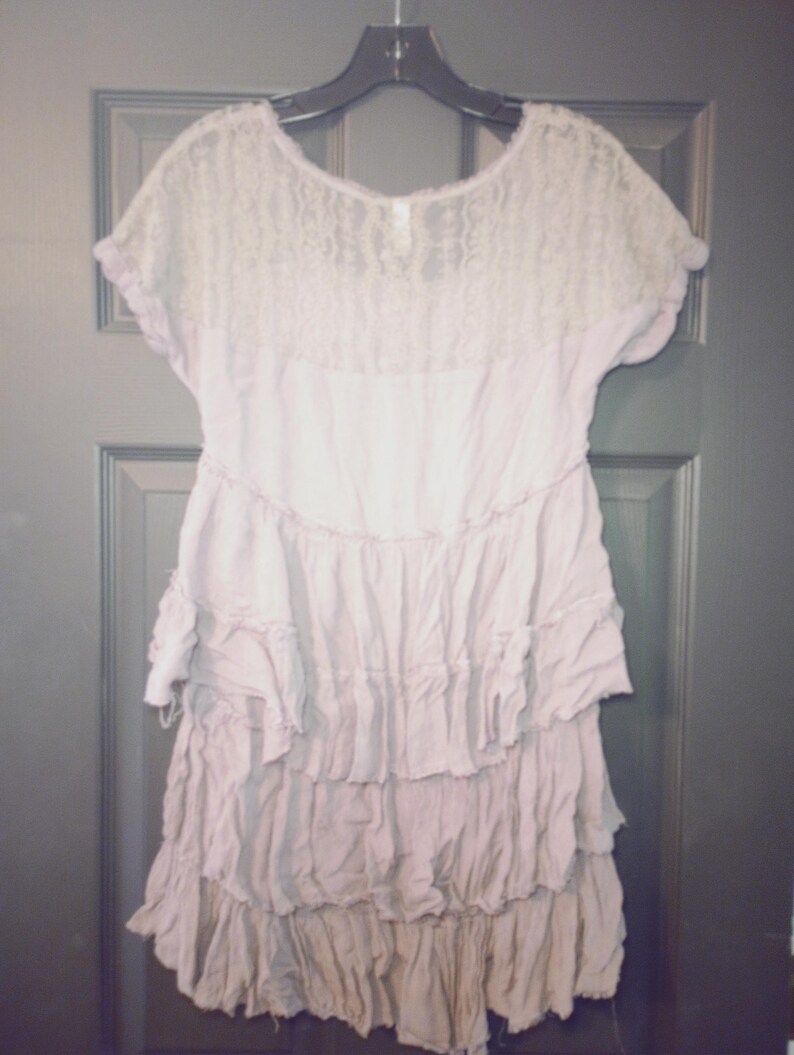 May include: A light purple dress with a lace bodice and short sleeves. The dress features a tiered, ruffled skirt. The garment is hanging on a black hanger against a dark gray door.