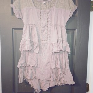 May include: A dusty rose-colored dress with short sleeves and a tiered, ruffled skirt. The yoke and sleeves are a light beige lace. The dress has embroidered details down the front and is hanging on a black hanger.