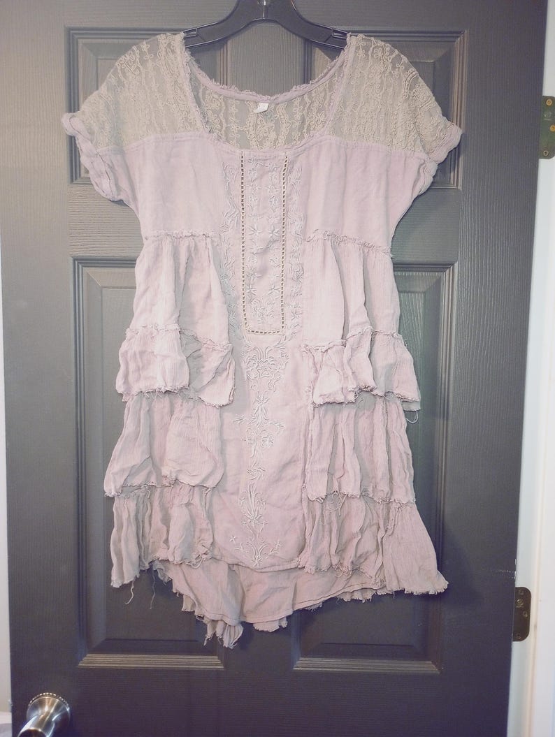 May include: A dusty rose-colored dress with short sleeves and a tiered, ruffled skirt. The yoke and sleeves are a light beige lace. The dress has embroidered details down the front and is hanging on a black hanger.