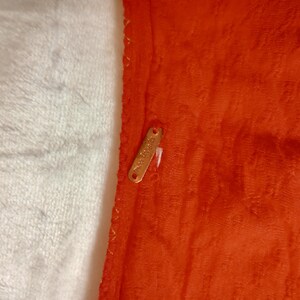 Puede incluir: Primer plano de una secci&oacute;n de tela naranja con una apariencia texturizada y afelpada. Una peque&ntilde;a etiqueta dorada est&aacute; adherida a la tela. La tela est&aacute; junto a un material blanco y esponjoso.