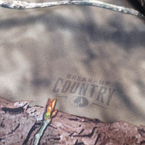 Puede incluir: Primer plano de una tela de camuflaje con las palabras "BREAK-UP COUNTRY" impresas en gris oscuro. La tela tiene un patr&oacute;n marr&oacute;n, verde y beige, con una peque&ntilde;a rama en primer plano.