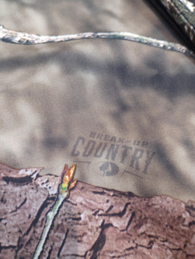 Puede incluir: Primer plano de una tela de camuflaje con las palabras "BREAK-UP COUNTRY" impresas en gris oscuro. La tela tiene un patr&oacute;n marr&oacute;n, verde y beige, con una peque&ntilde;a rama en primer plano.