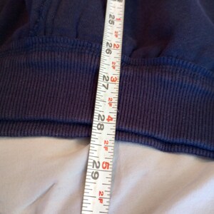 May include: Close-up of a navy blue sweatshirt with a ribbed hem. A white measuring tape shows a length of approximately 23 to 30 inches. The sweatshirt has a white stripe detail.