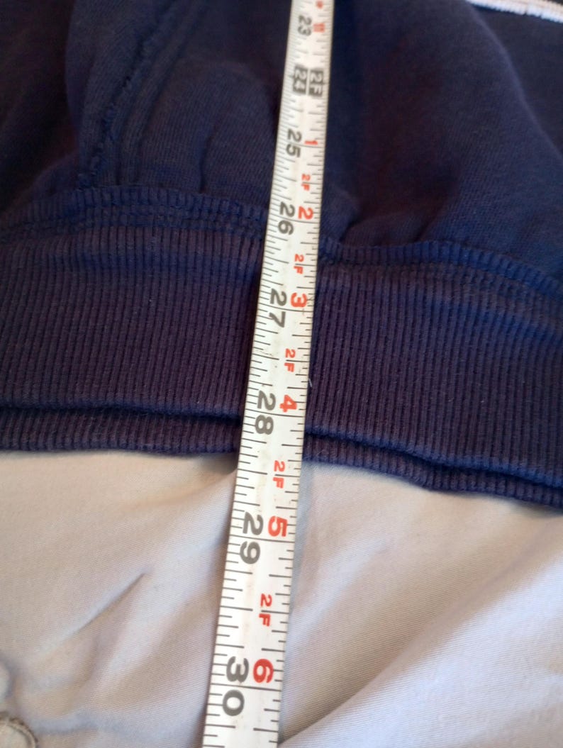 May include: Close-up of a navy blue sweatshirt with a ribbed hem. A white measuring tape shows a length of approximately 23 to 30 inches. The sweatshirt has a white stripe detail.