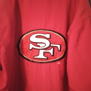 May include: Red jacket with a large embroidered San Francisco 49ers logo on the back. The logo is in white and gold with a black outline. The jacket has a hood and a zipper closure. The text "TM" is below the logo.