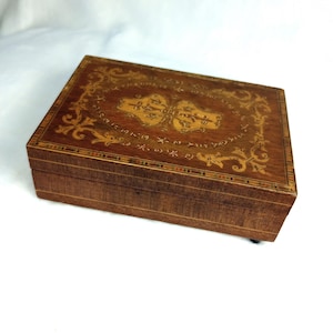 May include: A rectangular wooden box featuring intricate marquetry inlay on the lid. The lid displays a central light-colored wood floral motif surrounded by a darker wood border with scrolling leaf patterns and a multi-colored geometric edge.