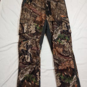 Puede incluir: Pantalones cargo de camuflaje con un patr&oacute;n marr&oacute;n, verde y beige. Los pantalones tienen m&uacute;ltiples bolsillos, cierre de bot&oacute;n y cintura y paneles laterales de color gris oscuro. La palabra "HART" est&aacute; impresa en naranja en la pierna derecha.