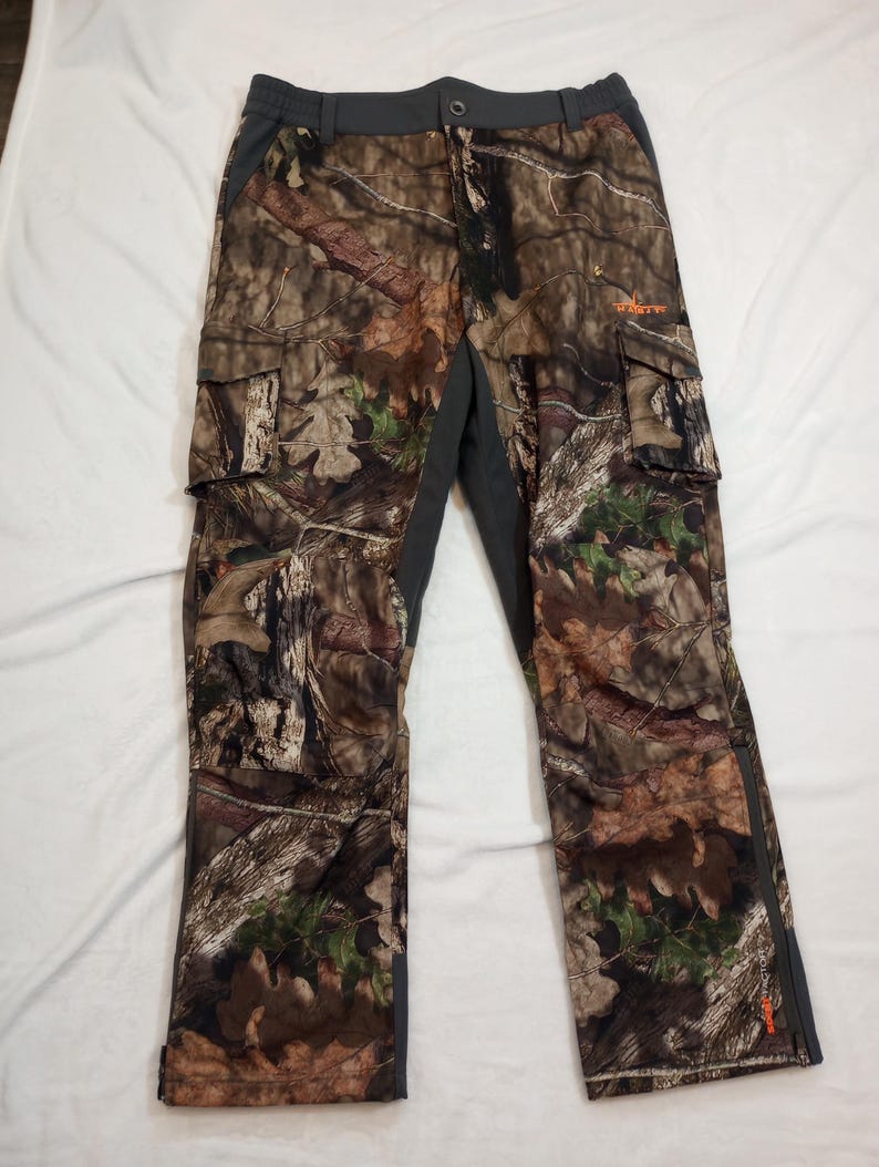 Puede incluir: Pantalones cargo de camuflaje con un patr&oacute;n marr&oacute;n, verde y beige. Los pantalones tienen m&uacute;ltiples bolsillos, cierre de bot&oacute;n y cintura y paneles laterales de color gris oscuro. La palabra "HART" est&aacute; impresa en naranja en la pierna derecha.