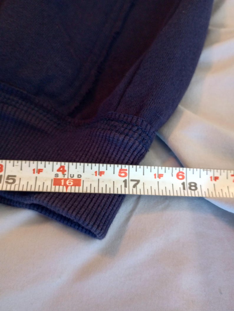 May include: Close-up of a navy blue sweatshirt cuff with a ribbed knit design. A measuring tape shows the cuff is approximately 6 inches wide. The fabric appears soft and slightly worn.