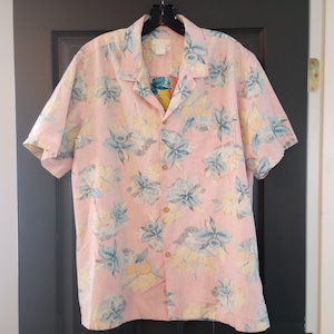May include: A short-sleeved button-down shirt in a light pink hue, adorned with a floral pattern in shades of blue and yellow. The shirt features a classic collar and wooden buttons. The shirt is hanging on a black hanger.