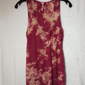 May include: A sleeveless burgundy vest top with a tie-dye pattern in beige and burgundy. It features a scoop neck and a button-up placket. The top is hanging on a black hanger.
