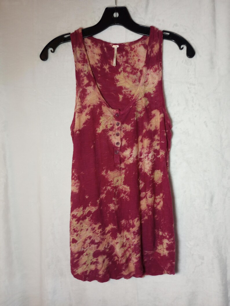 May include: A sleeveless burgundy vest top with a tie-dye pattern in beige and burgundy. It features a scoop neck and a button-up placket. The top is hanging on a black hanger.