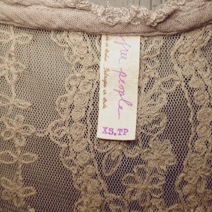 May include: Close-up of a beige lace top with a delicate floral pattern. The top features a scoop neckline and a tag that reads "Free People" in purple script, with "XS, TP" printed below.