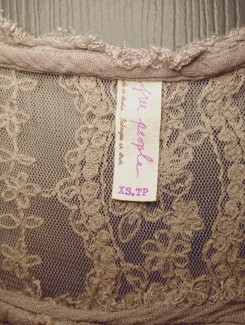 May include: Close-up of a beige lace top with a delicate floral pattern. The top features a scoop neckline and a tag that reads "Free People" in purple script, with "XS, TP" printed below.