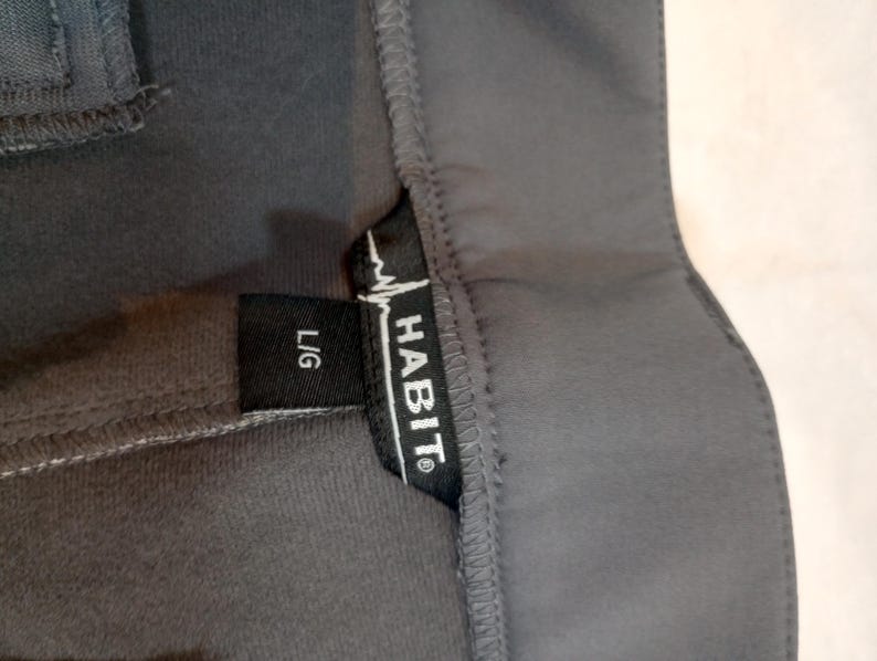 Puede incluir: Primer plano de una prenda gris oscuro con una etiqueta negra. La etiqueta dice "HABIT" en letras may&uacute;sculas blancas con un gr&aacute;fico de latido card&iacute;aco y "L/G" en blanco. La prenda parece estar hecha de un material suave y flexible.