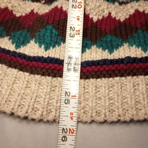 May include: A close-up of a beige knitted sweater with a brown, red, and green patterned design. A measuring tape is placed next to the sweater, showing a measurement of 24 inches.