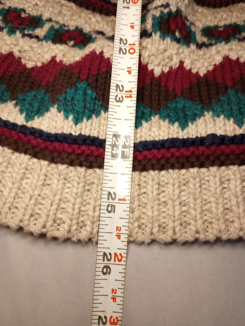 May include: A close-up of a beige knitted sweater with a brown, red, and green patterned design. A measuring tape is placed next to the sweater, showing a measurement of 24 inches.
