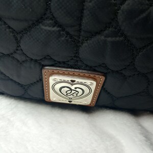 May include: Black quilted bag with heart-shaped stitching. The bag features a rectangular tag with a silver emblem and a brown leather border. The emblem has a heart design with the letters "B" and "S" intertwined.