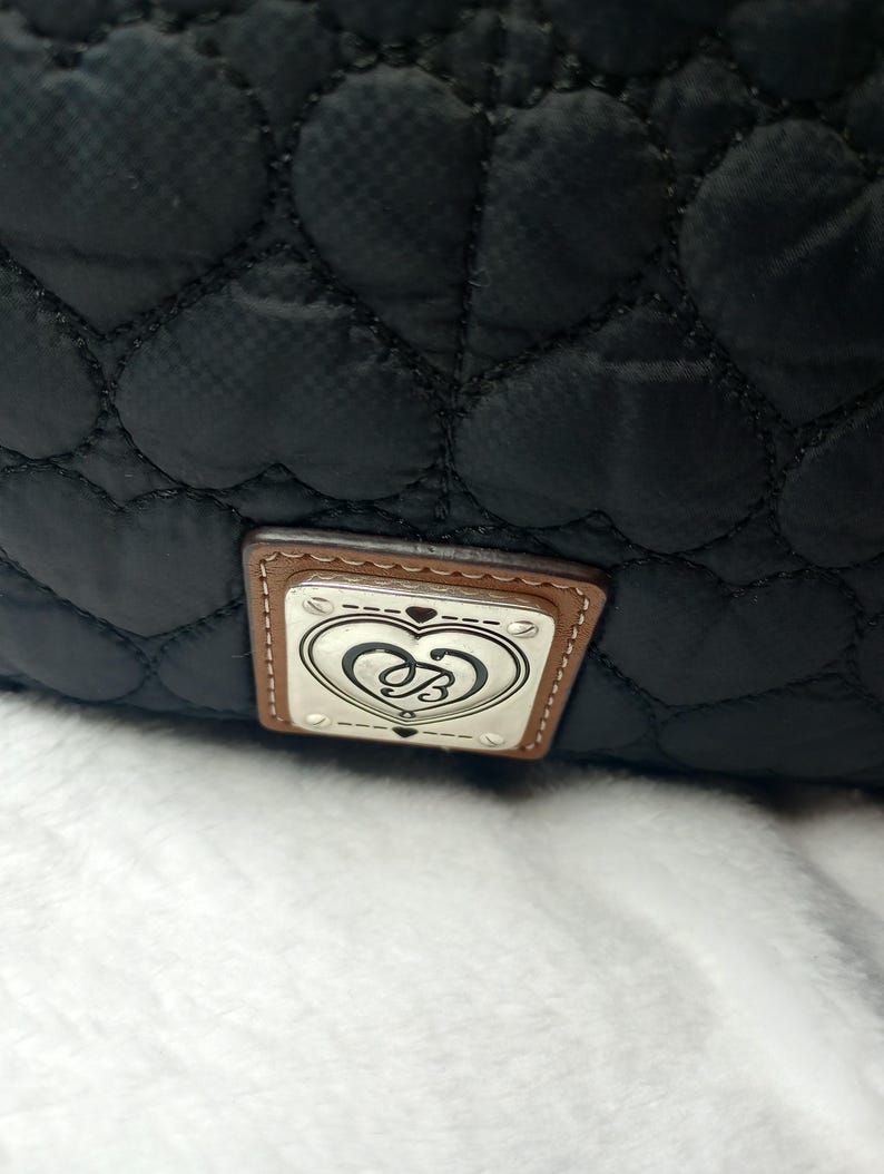 May include: Black quilted bag with heart-shaped stitching. The bag features a rectangular tag with a silver emblem and a brown leather border. The emblem has a heart design with the letters "B" and "S" intertwined.