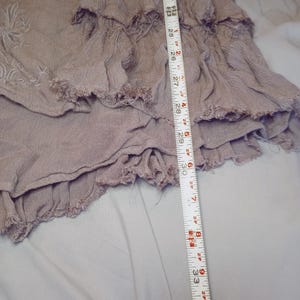 May include: A dusty rose-colored garment with layered ruffles and a frayed hem. The garment is measured with a tape measure, showing a length of approximately 34 inches. The fabric appears to be lightweight and textured, with embroidered details visible.