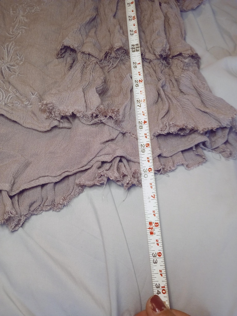 May include: A dusty rose-colored garment with layered ruffles and a frayed hem. The garment is measured with a tape measure, showing a length of approximately 34 inches. The fabric appears to be lightweight and textured, with embroidered details visible.
