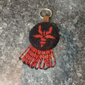 Könnte beinhalten: Ein Schlüsselanhänger mit Perlenbesatz und kreisförmigem Design. Der Schlüsselanhänger hat einen schwarzen Perlenhintergrund mit einem roten Sternmuster in der Mitte. Rote Perlen bilden einen Fransen am unteren Rand. Ein silberner Ring und ein brauner Lederriemen sind befestigt.