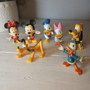 Puede incluir: Una colección de figuras de personajes de Disney, incluyendo Mickey Mouse, Minnie Mouse, Donald Duck, Daisy Duck y Pluto. Las figuras son de colores brillantes y posan en varias posturas. Las figuras son de plástico y se exhiben sobre una superficie blanca.