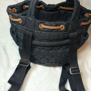 May include: Black quilted backpack with brown leather accents. Features a drawstring closure, top handle, and adjustable black shoulder straps. The bag has a heart-shaped quilted pattern.