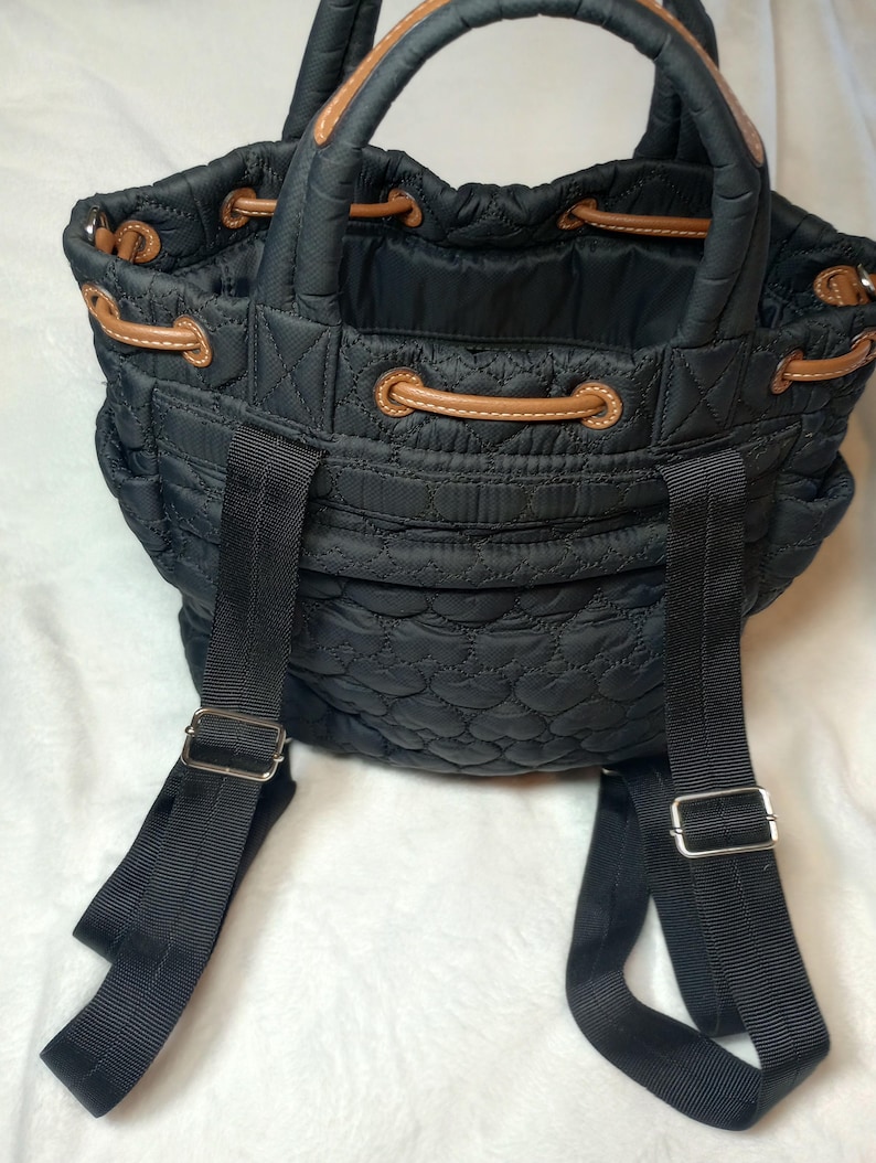 May include: Black quilted backpack with brown leather accents. Features a drawstring closure, top handle, and adjustable black shoulder straps. The bag has a heart-shaped quilted pattern.