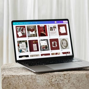 May include: A laptop displaying a website with various design templates. The screen shows a grid of images with a burgundy and white color scheme. The laptop is on a textured, light-colored stone surface.