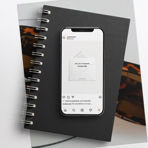 K&ouml;nnte beinhalten: Ein Smartphone zeigt einen Instagram-Post mit einem wei&szlig;en Umschlag und dem Text "20% Rabatt auf den gesamten Shop" und "Der Countdown l&auml;uft..."  Der Beitrag stammt vom Account "studioseven", und das Bild befindet sich auf einem schwarzen Notizbuch mit Spiralbindung.