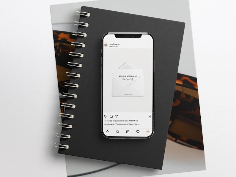 K&ouml;nnte beinhalten: Ein Smartphone zeigt einen Instagram-Post mit einem wei&szlig;en Umschlag und dem Text "20% Rabatt auf den gesamten Shop" und "Der Countdown l&auml;uft..."  Der Beitrag stammt vom Account "studioseven", und das Bild befindet sich auf einem schwarzen Notizbuch mit Spiralbindung.