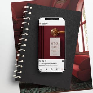 May include: A smartphone displays an "Out of Office" door hanger on a burgundy door. The phone rests on a black spiral-bound notebook. The background includes a red sofa and a window.