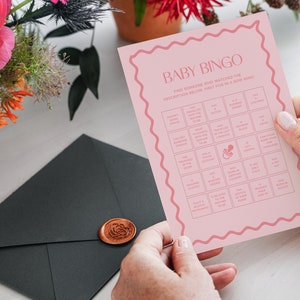May include: Pink baby bingo card with a pacifier icon in the center. The card has a grid of squares with various descriptions for guests to find someone who matches the description. The text on the card reads "Baby Bingo Find someone who matched the description below. First five in a row wins!"