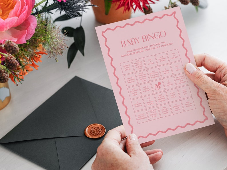 May include: Pink baby bingo card with a pacifier icon in the center. The card has a grid of squares with various descriptions for guests to find someone who matches the description. The text on the card reads "Baby Bingo Find someone who matched the description below. First five in a row wins!"