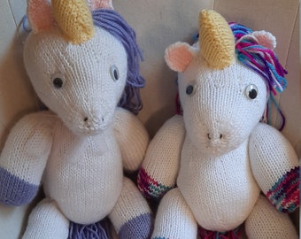 May include: Two white knitted unicorn toys with colorful yarn tails and horns. The unicorn on the left has a purple tail and horn, while the unicorn on the right has a pink, blue, and purple tail and a yellow horn.