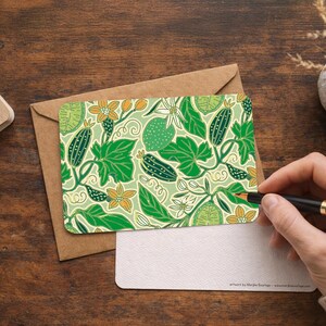 Può includere: Biglietto con motivo botanico verde e bianco con cetrioli, foglie e fiori. Il biglietto è su una busta marrone, con un biglietto bianco vuoto e una penna. Opera d'arte di Morpha Boutique.