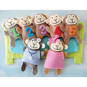 May include: Five brown felt finger puppets shaped like monkeys, each with a different number on their chest. The numbers are 1, 2, 3, 4, and 5. There are two additional felt puppets, one wearing a blue doctor's coat and the other wearing a pink dress. All the puppets are sitting on a green and blue felt bed.