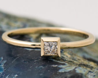 May include: A gold ring with a square-cut diamond. The ring has a slender, polished band and a bezel setting. The square gemstone is the focal point. The ring is presented on a textured, blue-green surface.