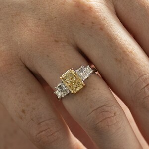 May include: A gold ring with a large, rectangular, yellow diamond in the center. The ring has two smaller, square, white diamonds on either side of the center stone.