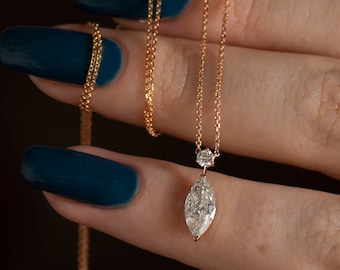 May include: A delicate gold chain necklace featuring a marquise-cut diamond pendant suspended from a small round brilliant-cut diamond accent stone. The fine metal chain is crafted from a warm-toned gold, showcasing a minimalist and elegant design.
