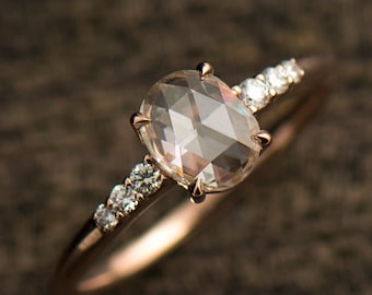 May include: A rose gold engagement ring with a large, oval-shaped, clear, faceted gemstone surrounded by a band of smaller, clear, round gemstones.