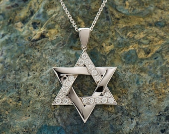May include: A silver pendant necklace with a diamond-encrusted Star of David charm. The pendant is on a rocky surface.