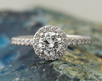 May include: A silver engagement ring with a round, clear diamond center stone surrounded by a halo of smaller diamonds. The ring is set on a textured, blue and green surface.