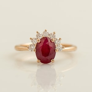 Puede incluir: Un anillo de oro con una gran piedra de rubí ovalada rodeada por un halo de diamantes más pequeños.