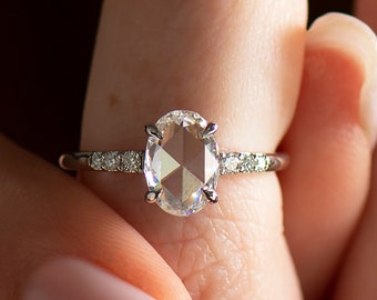 May include: A silver ring with a large, clear, oval-shaped diamond. The diamond has a unique, faceted cut. The ring has a thin band with smaller diamonds set along the sides.