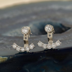 May include: A pair of silver-toned earrings. Each earring features a large, round, clear stone at the top and a curved row of smaller, clear stones. The earrings are designed to be worn with the curved row of stones facing downward.