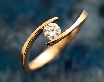 Op de afbeelding: Een gouden ring met een enkele, ronde, heldere diamant gezet in een uniek, gebogen ontwerp.