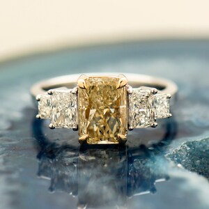 May include: A gold and white engagement ring with a large, rectangular, yellow diamond in the center and two smaller, square, white diamonds on either side.